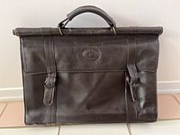 Single dark brown leather briefcase with dual buckle front flap and top handle, Pierre Cardin logo embossed in center front.
