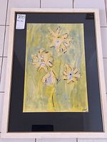 Framed watercolor painting showing four yellow flowers with black outlines on a greenish-yellow background, with signature visible, mat and frame also visible