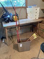 Two archery bows and six arrows standing in a metal pail, set on garage floor next to workbench.