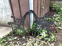 Two black cast iron bench frames standing against a wooden shed, showing the front design with floral and scrollwork details among garden plants.