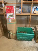 Unopened boxed artificial Christmas tree and green bin with assorted Christmas lights in garage