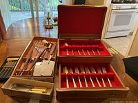 Photo showing open wood cutlery case with red interior and Tudor stainless steel forks and spoons arranged inside. Barbecue tools with wooden handles next to case on table.