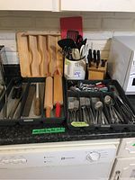 Photo of cutlery organizer with spoons, forks, knives and large kitchen utensils, wooden rolling pins and wooden cutting block in background.