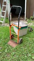 Orange metal dolly cart with black handle grips and two wheels standing on grass in front of a shed.