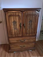 Front view of tall wood veneer dresser, showing carved floral doors and two metal-pulled drawers.