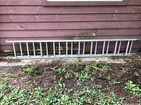 Full length view of aluminum extension ladder laying horizontally against a wood siding wall on ground covered with grass and dirt.