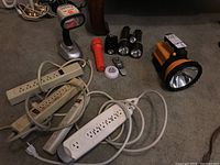 Photo showing five flashlights, one portable lamp, four power bars, a remote control, and a round white button-like device all placed on floor.