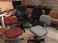 Four adjustable office chairs including one black faux leather chair with armrests and three fabric upholstered chairs in black, light gray, and rust colors.