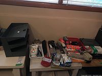Full view of assembled desk items including a black metal three-tier shelf, multiple staplers, assorted writing instruments, packs of staples, boxes of paper clips, a tape dispenser, rulers, and containers of Brother tape and other office supplies.