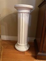 White ceramic plant stand with fluted column design, standing on wooden floor next to furniture