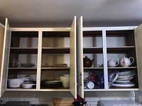 Full view of two open kitchen cupboards showing stacked plates, bowls, teapots, disposable cups, plates, and utensils.