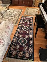 Photo displaying the maroon and dark blue geometric patterned runner rug with fringes beside a piano and floral sofa on a wooden floor