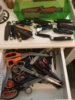 Image showing variety of vintage folding cutters and assorted scissors packed on a media storage container and drawers. Some cutters have worn and vintage appearance with mixed handle materials and colors. Scissors visible in various sizes and handle types including plastic and metal.