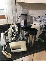Hamilton Beach food processor, hand mixers, electric cutter, thermos, and travel cup pictured on kitchen counter with cords tangled
