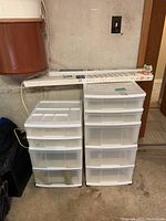 Front view of two white plastic storage containers, one with multiple drawers and the other with drawers and top compartment, along with two window blinds in packaging placed on top.