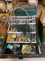 Tackle box open showing multiple tray compartments with various fishing lures and accessories including a fishing net on top.