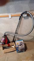 Red Dirt Devil Featherlite canister vacuum with hose and attachments, placed on floor and leaning against wall with a cardboard box of accessories.