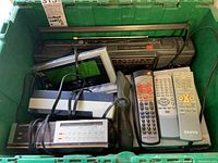 Green bin containing a large vintage radio, DVD player, battery charger, and several remote controls