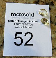 Two loose faceted oval citrine gemstones placed on a numbered auction lot card on a granite surface for scale and identification.