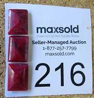 Three rectangular, faceted ruby gemstones displayed on a white card with auction information, showing the overall appearance and size relative to the card.