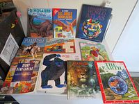 Photo of a collection of children's books showing a variety including dinosaur books, world atlas, storybook, and activity books on a white surface.