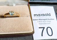 Front view of gold ring with central blue gemstone and clear side stones in beige cushioned ring box