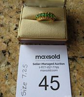 Front view of gold-tone ring with green gemstones and clear stones, displayed in a beige ring box with size 7.25 noted on auction card.