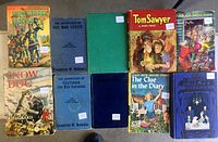 Nine vintage children's books arranged in two rows on a beige fabric. Covers show colorful illustrations including knights, dogs, and children. Titles include Saint George For England, The Adventures of Old Man Coyote, and Tom Sawyer.