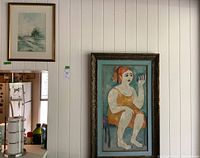 View of two framed paintings hanging on white paneled wall. Left is smaller framed watercolor landscape; right is larger textured oil painting of red-haired lady.