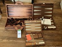 Three open backgammon and chess game sets shown on wooden floor. The first set has a velvet-like playing surface with backgammon points, dice cups, dice, and playing pieces. The second set is an ornately carved wooden box with wooden backgammon pieces scattered inside. The third image shows a worn brown leather carry case that holds the portable chess and backgammon combo game.