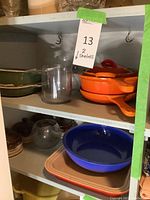 Two-shelf overview showing green and white enamel dishes, orange Descoware cookware, glass vases, brown bowls, blue and peach bakeware