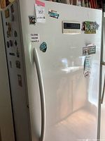 Front side view showing white freezer with handle and electronic control panel on door, covered in fridge magnets and stickers.