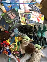 Photo showing the assortment of children's books with colorful covers, toys including Baby Yoda dolls, Transformers figure, and various plush animals, along with some children's shoes in the background.