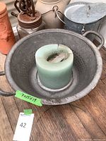 Large metal colander with worn surface and green tape showing dimensions inside, placed on wooden floor with other pottery items in background.