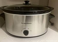 Front view of Hamilton Beach 33276 Slow Cooker showing stainless steel body, black handles, glass lid with knob, and control knob.