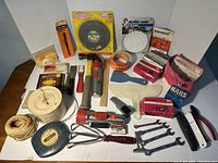 Photo of a varied assortment of vintage hand tools and hardware including tape measures, nails, scrapers, wire brush, wrenches, and a caulking gun arranged on a table surface.
