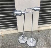 Pair of vintage silver floor lamps shown outdoors with a wall and shutter backdrop. Both lamps have round bases, tall poles, curved arms, and oval lamp heads. One lamp's cord is visible coiled on the floor.