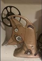 Side view showing overall projector with belt drive and film reel arm, bronze textured finish.