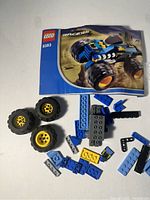 Instruction manual laid on a table next to four large LEGO wheels with yellow rims and several blue, black, and gray LEGO pieces scattered around.