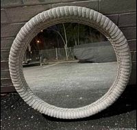 Front view of the large round mirror with white wicker frame leaning against a brick wall outdoors at night.
