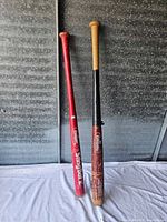 Two baseball bats; one red, one black with natural handle, against a metal wall background