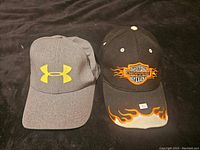 Both baseball caps shown side by side on a dark fabric background. The Under Armour cap is grey with a yellow logo, the Motor Choppers cap is black with flame decoration.