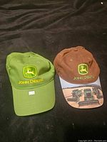 Two John Deere branded baseball caps: a plain green one and a brown one with a tractor image printed on the brim.