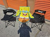 Three folding camping chairs, two black and one SpongeBob SquarePants themed, shown unfolded and leaning against an orange storage unit door.