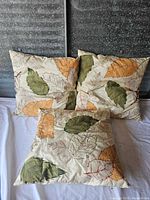 Three decorative pillows with multicolored leaf embroidery arranged on a white surface and dark textured background.