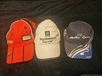 Three racing ball caps displayed together on black background: Dodge Stanley in orange/black, GM Goodwrench in beige, Norton Racing in dark gray/blue.