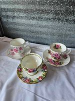 Three Paragon Tapestry Rose fine bone china teacup and saucer sets displayed on white cloth, showing the floral designs and variety of colors and trims.