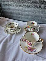 Three porcelain teacups each with distinct floral patterns and gold trim, paired with matching saucers arranged on fabric