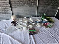 Full view of assorted cups, mugs, glassware including a glass bowl and decorative ceramic items on white cloth.