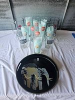Photo showing 11 assorted beer glasses grouped together with a black round serving tray in front featuring an elephant design.